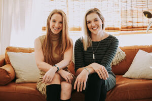 a portrait of 2 young smiling women on a couch in a bright indoor setting