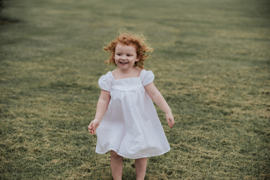 A cheerful young girl with curly red hair, wearing a white dress, playing and smiling in an open grassy field.