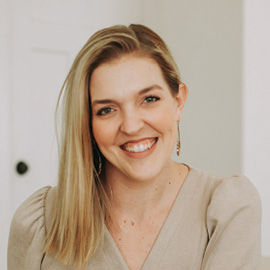 Portrait of a smiling woman with long blonde hair, wearing a beige top, captured in a bright indoor setting.